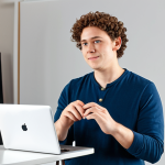 A professional musical theater artist, fully clothed in modest, appropriate audition attire, stands confidently in a natural pose, performing a virtual audition. The artist is a young adult, looking directly at a webcam on a laptop. The background is a clean, well-lit home studio setup with a neutral backdrop and a visible microphone.
    Quality: professional photography, high resolution, perfect anatomy, correct proportions, natural body proportions, well-formed hands, proper finger count, safe for work, appropriate content, fully clothed, family-friendly.